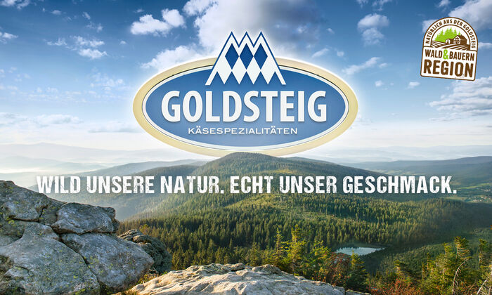 Startseite: Blick vom Arber in die Weite des sommerlichen Bayerischen Waldes. Mittig ist das GOLDSTEIG-Logo, darunter der Slogan "Wild unsere Natur. Echt unser Geschmack.“ zu erkennen.